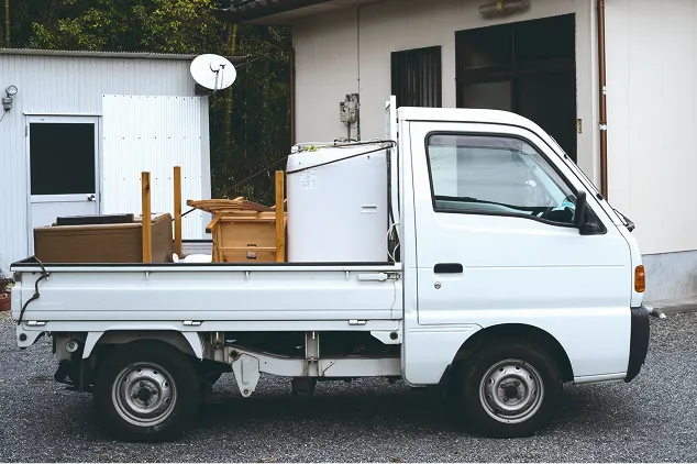 生活家電や家具などの荷物を荷台にたくさん積み込み、建物の前に停車している白い軽トラック