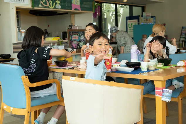 子ども食堂のテーブルを囲み、笑顔でカメラに向かって楽しそうに手を伸ばして遊ぶ子どもたち