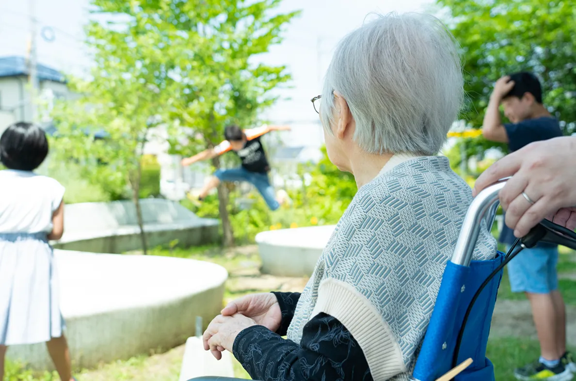 緑豊かな庭で元気に遊ぶ子どもたちを、車椅子から優しい眼差しで見守る高齢の利用者の後ろ姿