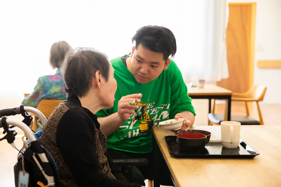 車椅子の利用者の目線に合わせ、優しく語りかけながら温かい雰囲気で食事の介助をおこなうスタッフ