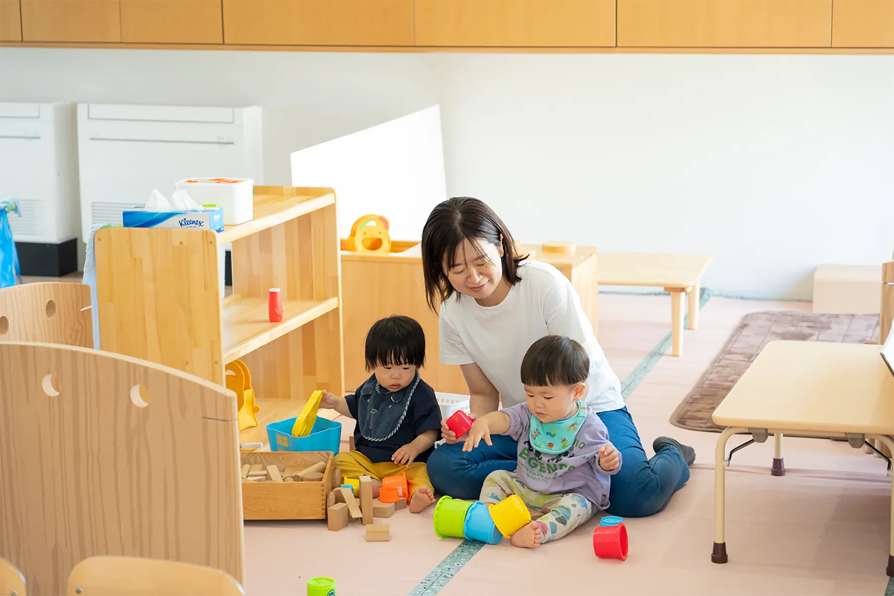 明るい保育室内で、床に座って小さなおもちゃで遊ぶ子どもたちを穏やかな笑顔で見守るスタッフ