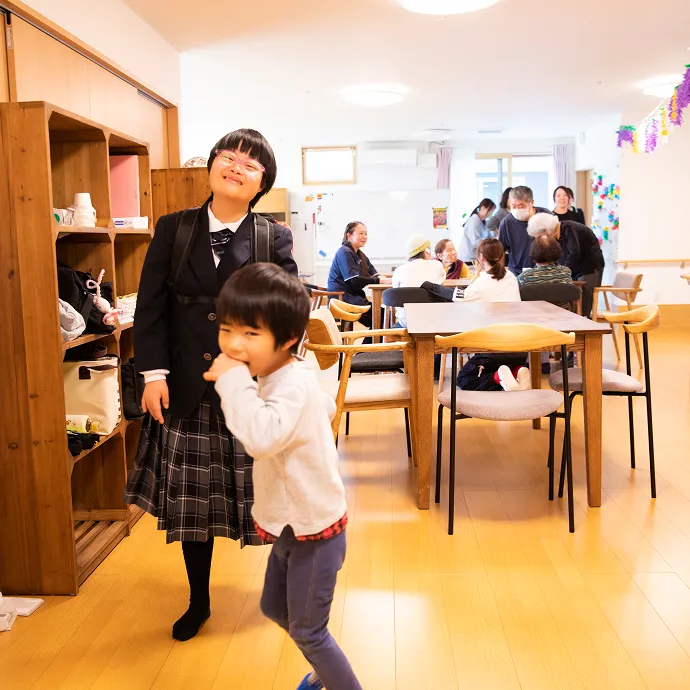 明るい施設内で、笑顔を見せる子どもたちと、奥のテーブルでスタッフや高齢者が談笑する多世代交流の風景