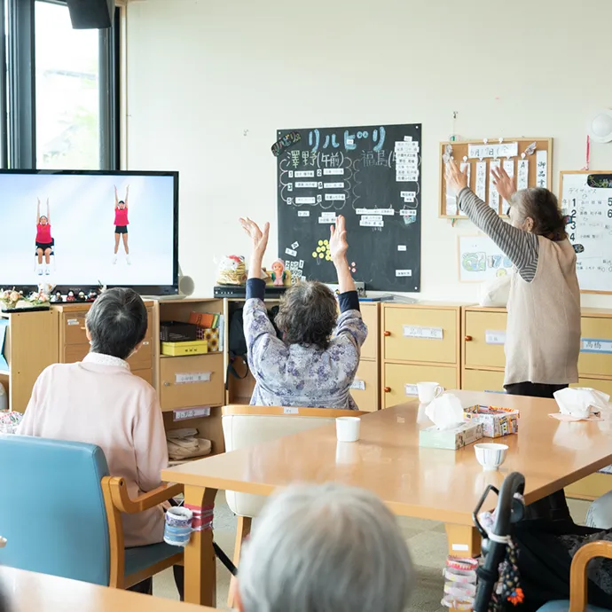 明るい室内のデイサービスで、テレビ画面の映像に合わせて両手を挙げ、体操を楽しむ高齢者たち