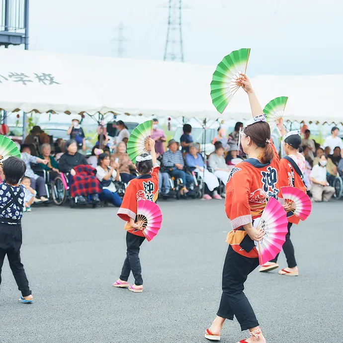 屋外の特設会場で、お揃いの法被を着て色鮮やかな扇子を手に踊りを披露する子どもや地域の人々