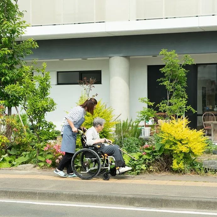 施設の前に広がる色鮮やかな花や緑の植栽の横を、スタッフに車椅子を押されて穏やかに散歩する高齢者