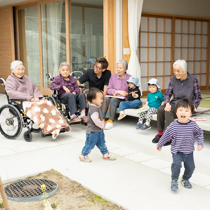 施設の縁側で、車椅子の高齢者やスタッフに見守られながら元気に遊ぶ小さな子どもたちの姿