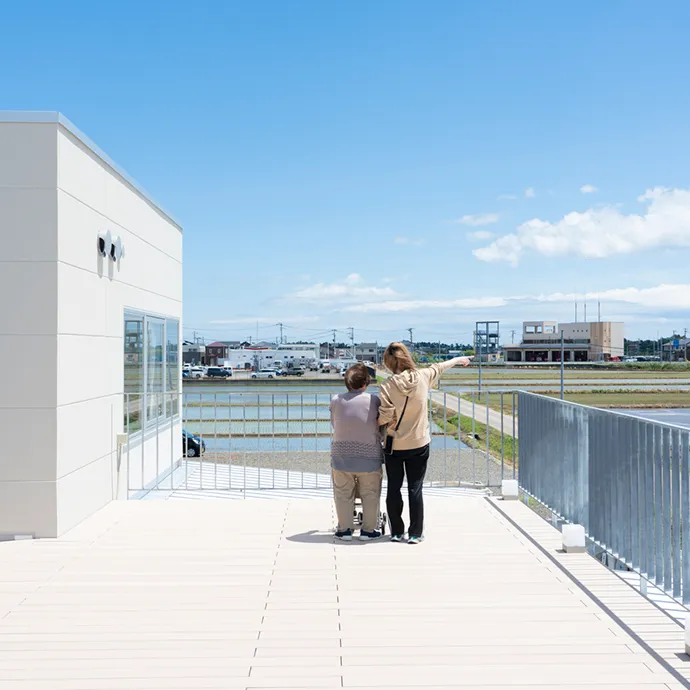 青空が広がる開放的な屋上テラスから、スタッフと一緒にのどかな田園風景を眺める高齢の利用者