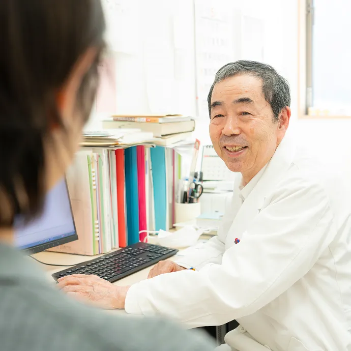 診察室のデスクで、患者の話に耳を傾けながら優しく温かい笑顔を見せる白衣の医師